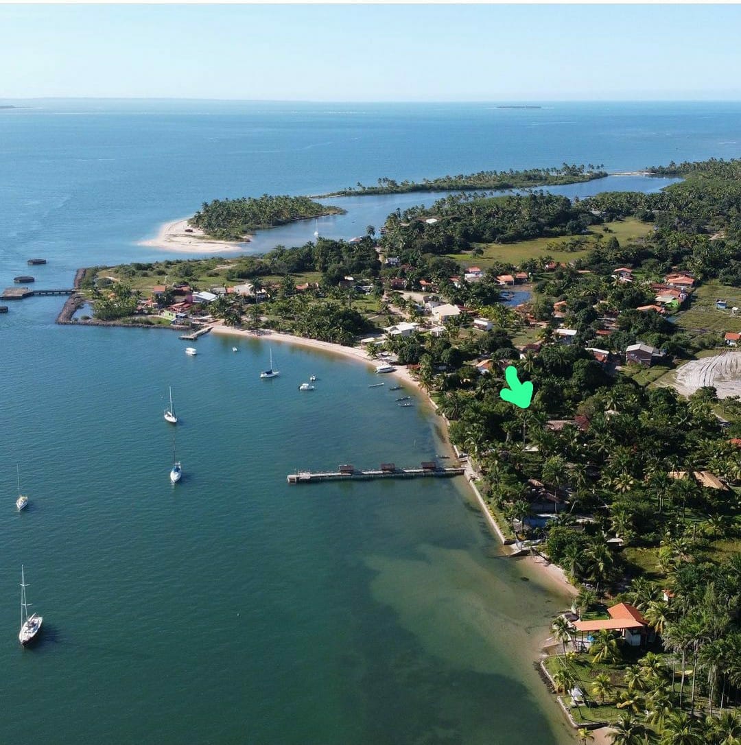 An aerial view captures a serene coastal landscape featuring a sandy beach alongside calm waters. Lush green foliage surrounds a collection of homes, while sailboats rest in the bay. The soft shoreline and nearby palm trees enhance the tranquil atmosphere.