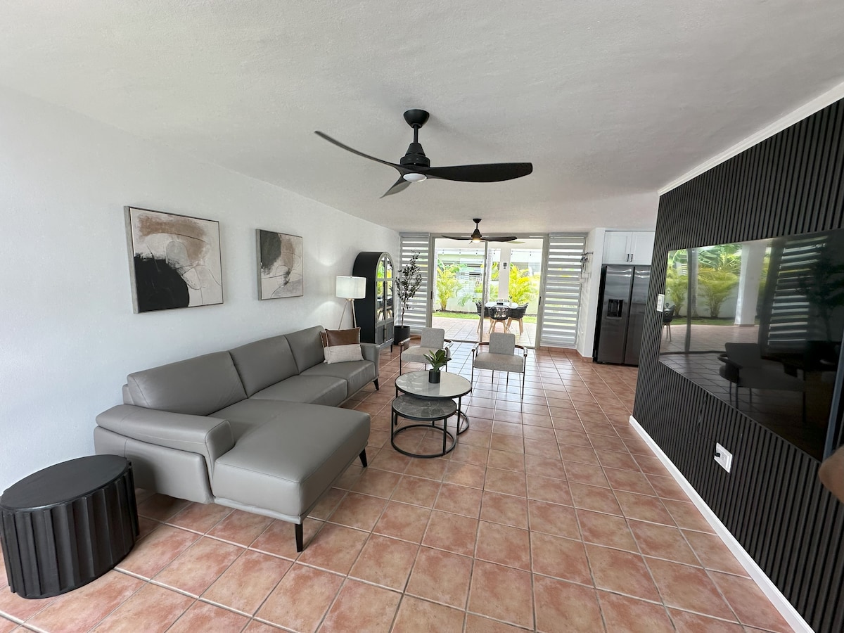 A modern living area features a light gray sectional sofa paired with a circular black table. A flat-screen TV is mounted on a textured wall. Natural light flows through large sliding doors, leading to an outdoor space filled with greenery.