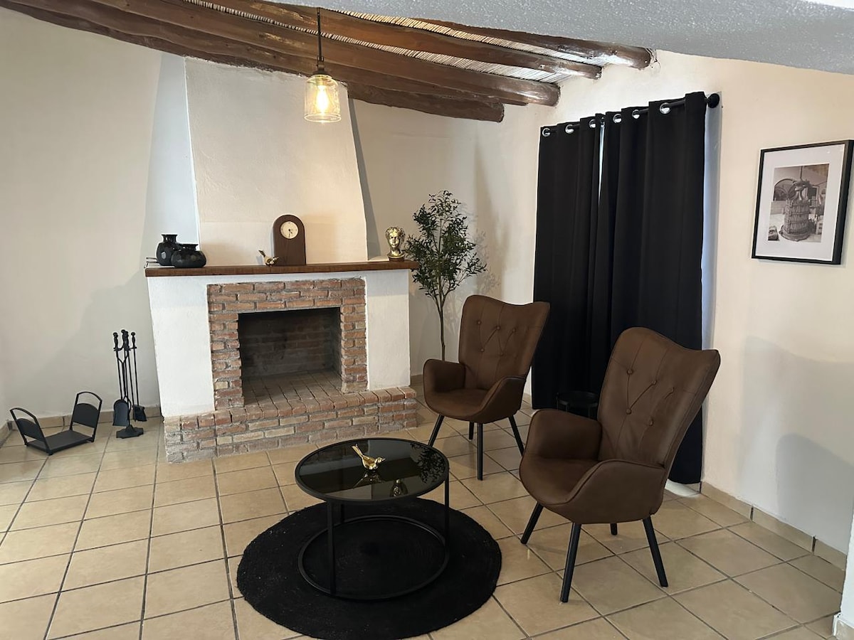 A cozy living area features two brown armchairs positioned beside a circular black coffee table. An exposed brick fireplace serves as a focal point, accompanied by decorative elements. The space is complemented by a small indoor plant and dark curtains that frame a window.