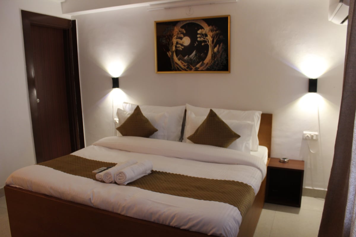 A spacious bed is centered in the room, adorned with two accent pillows. Soft lighting from wall-mounted fixtures highlights the bed and artwork above. A small bedside table is present on one side, and light-colored curtains complement the neutral-toned walls.