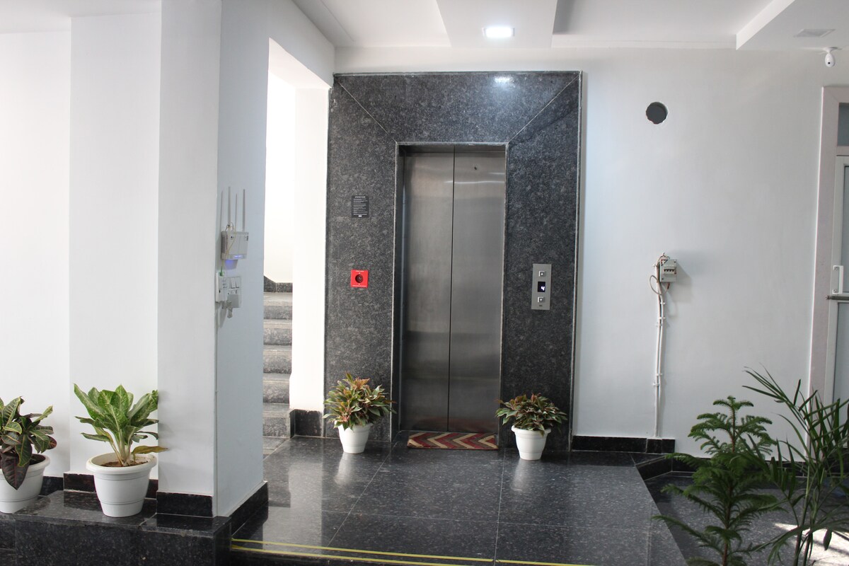 An elevator is positioned against a sleek, dark granite wall in a well-lit corridor. Potted plants are placed on either side of the elevator, enhancing the overall ambiance. Stairs are visible in the background, accessible for alternative means of ascent.