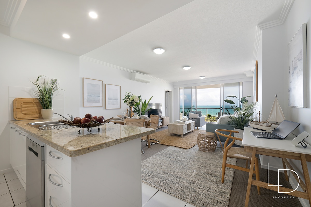 An open-plan kitchen and living area features a stone countertop with a fruit bowl, leading to comfortable seating arrangements. Large windows provide abundant natural light and showcase ocean views. A neutral color palette is complemented by decorative plants, creating a welcoming space.