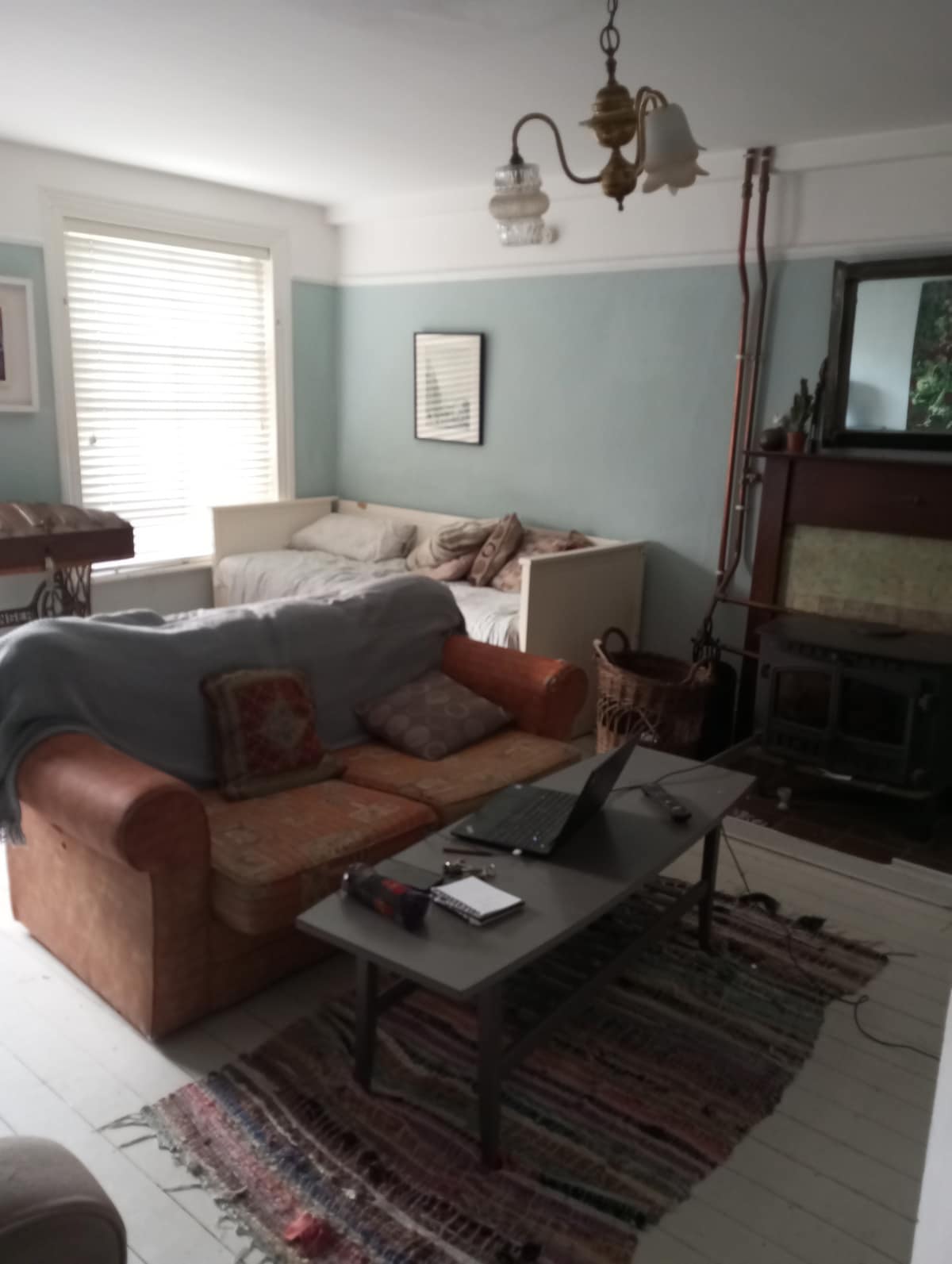 A comfortable living area features a sofa adorned with patterned cushions and a coffee table. Light is provided by a classic chandelier above. A cozy sleeping space is visible in the background, with a mix of natural lighting filtering through the window and illuminating the wooden floor.