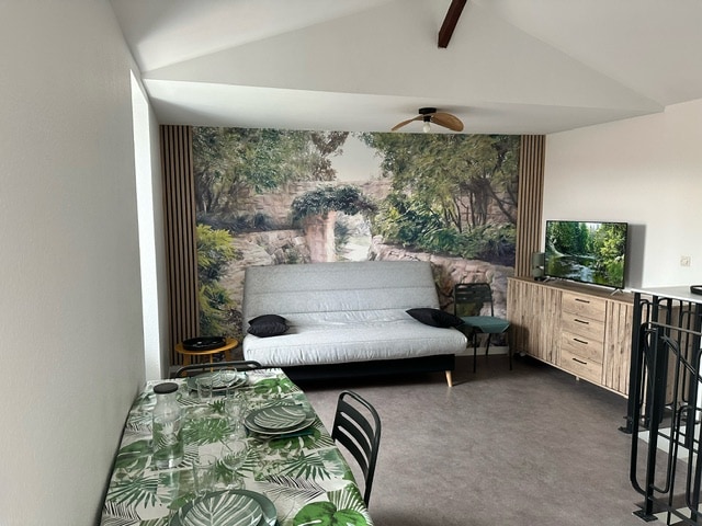 A spacious living area features a comfortable sofa against a backdrop of a nature-inspired mural. A dining table is set with dinnerware on a patterned tablecloth, and a television stands on a modern sideboard. Natural light illuminates the room from the windows above.