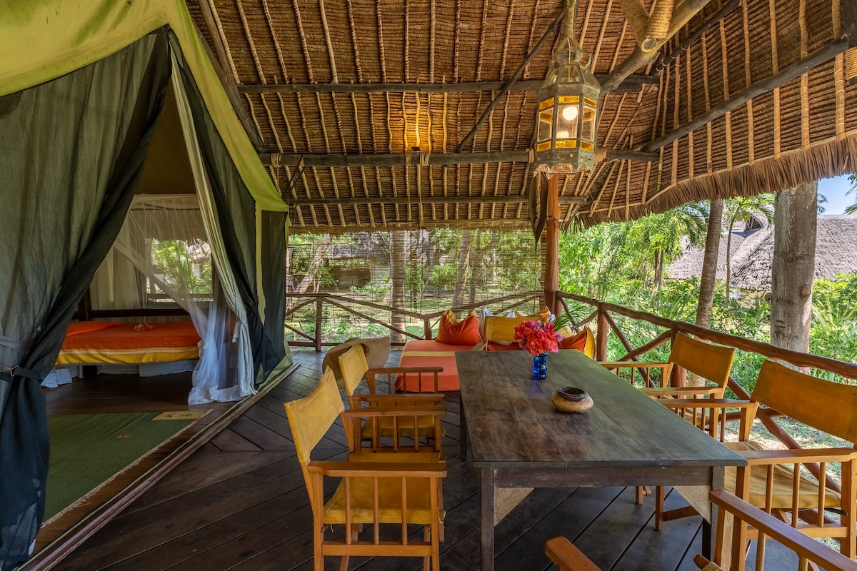 An outdoor seating area features a wooden table surrounded by several chairs. A lantern hangs from the ceiling, providing soft illumination. A safari-style tent is visible, with a spacious bed inside. Lush greenery is seen in the background, enhancing the tropical setting.