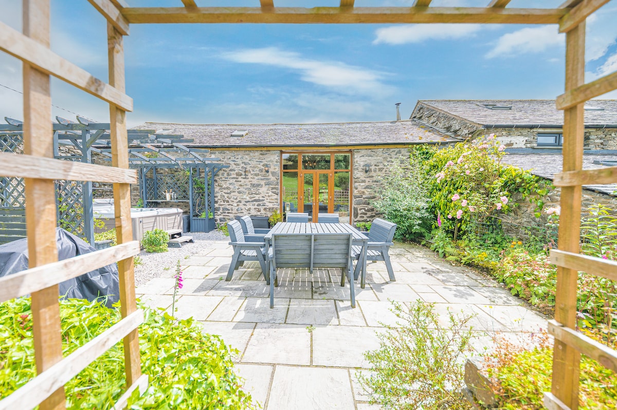 An outdoor terrace is presented with a stone-paved area, featuring a large dining table surrounded by six chairs. Lush greenery and climbing plants frame the space, while a hot tub is visible in the corner. The rustic barn structure forms the backdrop.
