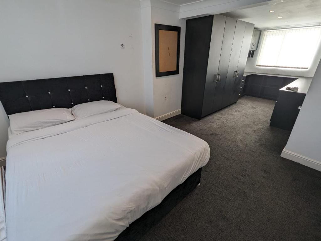 A bright bedroom features a spacious bed with a neatly made white comforter. A dark wardrobe with multiple doors is positioned along one wall, while a work desk and chair are visible near the window, which is dressed with light, sheer curtains.