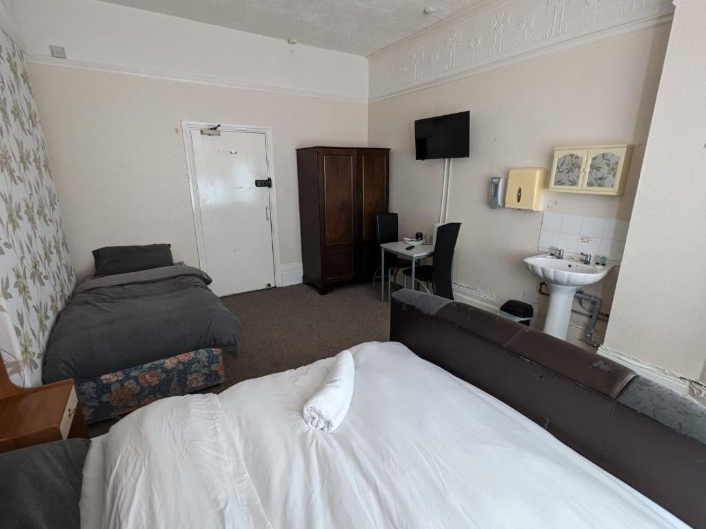 A well-organized room features two sleeping areas with simple bedding and ample carpeted space. A compact desk with a chair and a sink are visible, while a wall-mounted television provides entertainment options. Light-colored walls and a floral accent wall add a touch of decor.