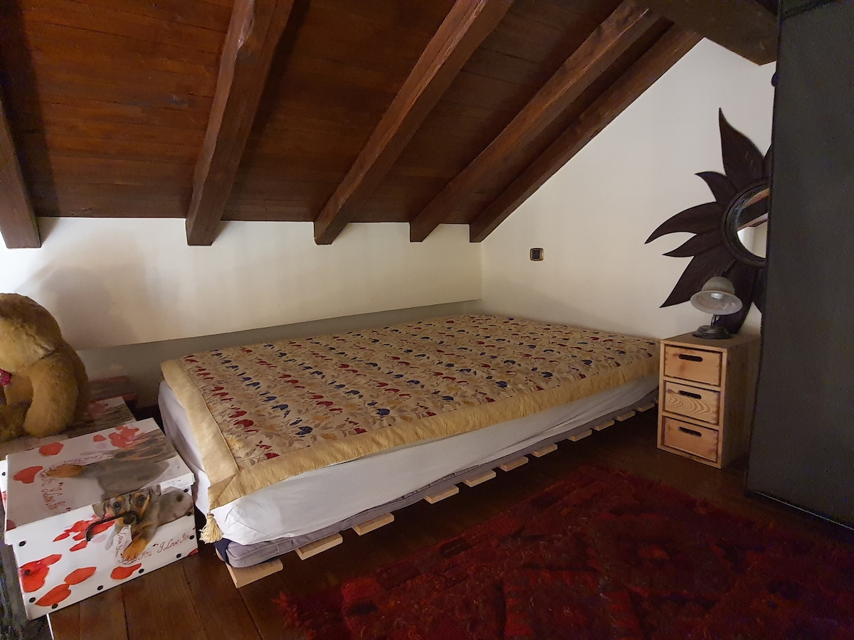 A cozy sleeping area is defined by a spacious bed adorned with a patterned quilt. Wooden beams create a warm ceiling above. A bedside lamp and a small dresser complement the space, while a plush bear sits nearby, enhancing the inviting ambiance.