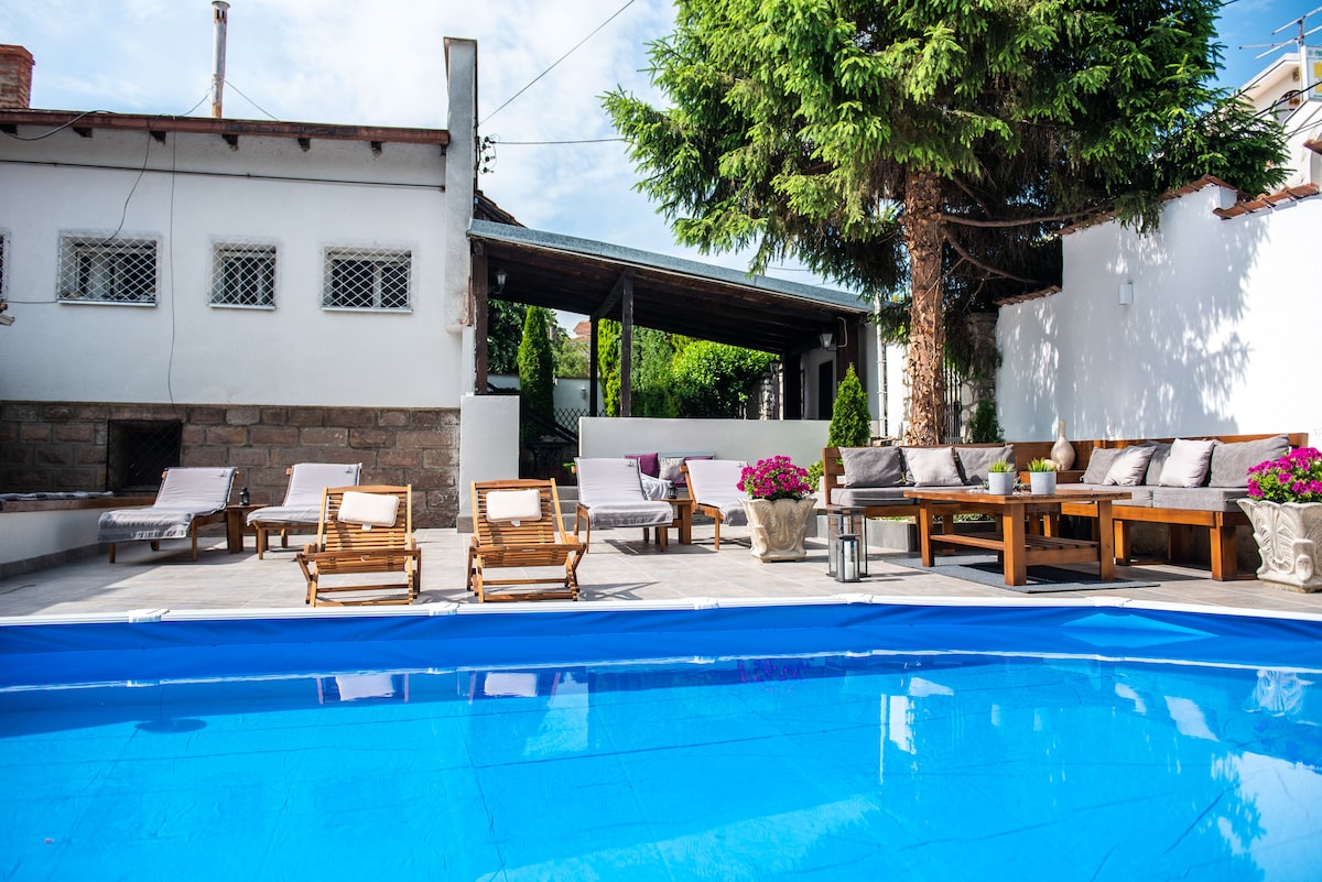 A private pool glistens in the foreground, framed by comfortable lounge chairs and shaded seating areas. The background features a spacious terrace with wooden furniture and vibrant flowers, surrounded by lush greenery and a welcoming atmosphere.