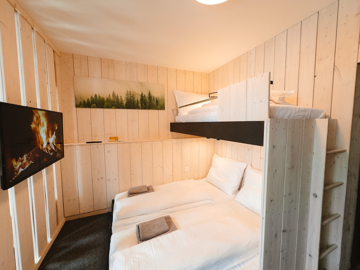 A modern room is presented with wooden paneling, featuring a spacious double bed and a lofted single bed. A television is mounted on the wall opposite the beds. Soft gray towels are neatly arranged on each bed, and a calming forest scene is displayed above.