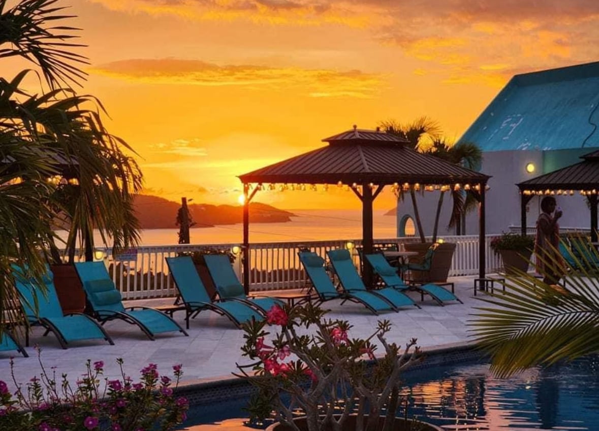 Lounge chairs in vibrant teal are arranged around a pool area, offering views of a sunset over the water. Tropical plants and flowers are visible in the foreground, with a structure featuring a covered area in the background, enhancing the serene ambiance.