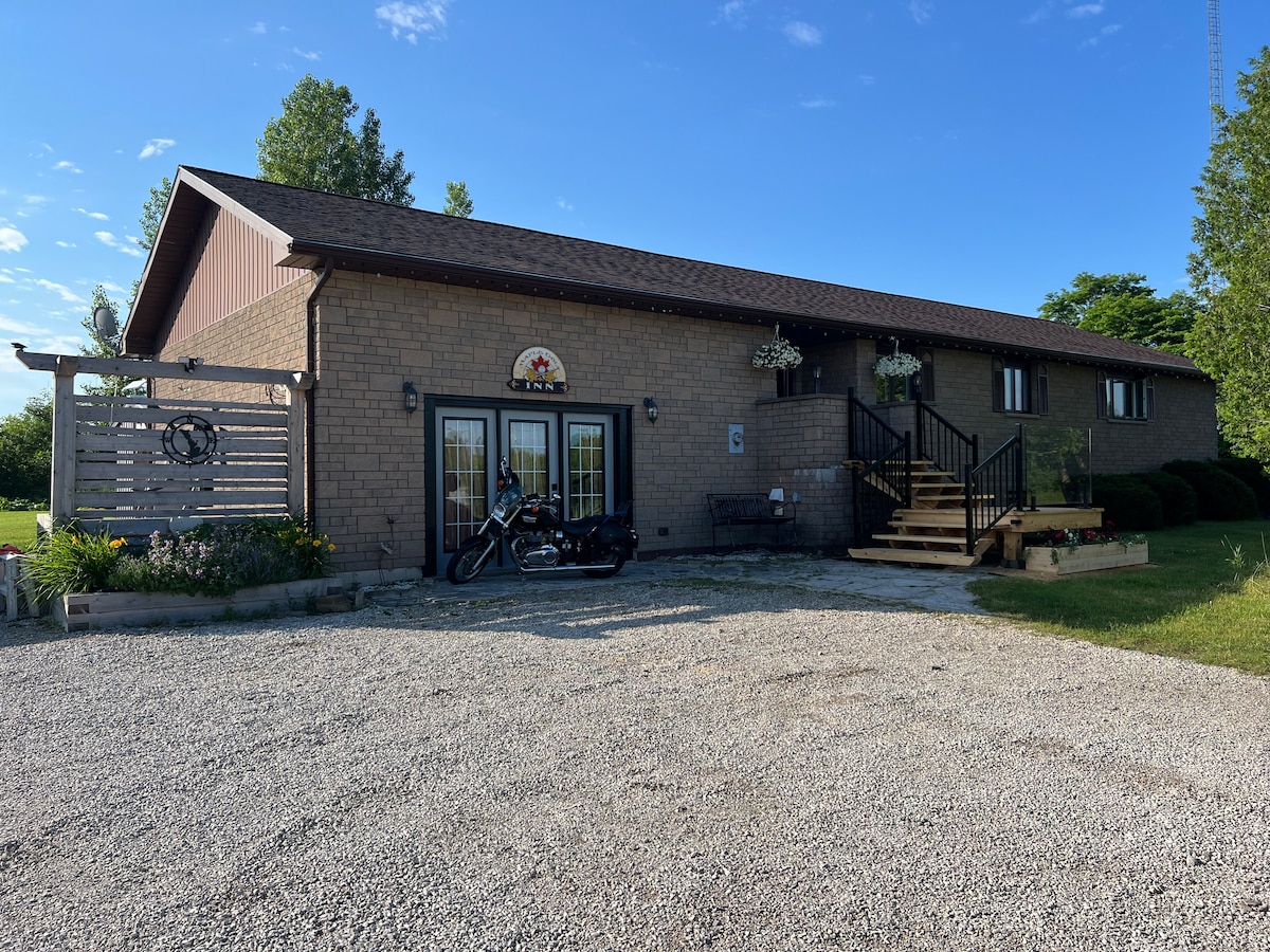 A spacious brick home is set against a clear blue sky, featuring an inviting entrance with steps leading up to double glass doors. Flower planters adorn the exterior, and a motorcycle is parked nearby, enhancing the approachable countryside ambiance.