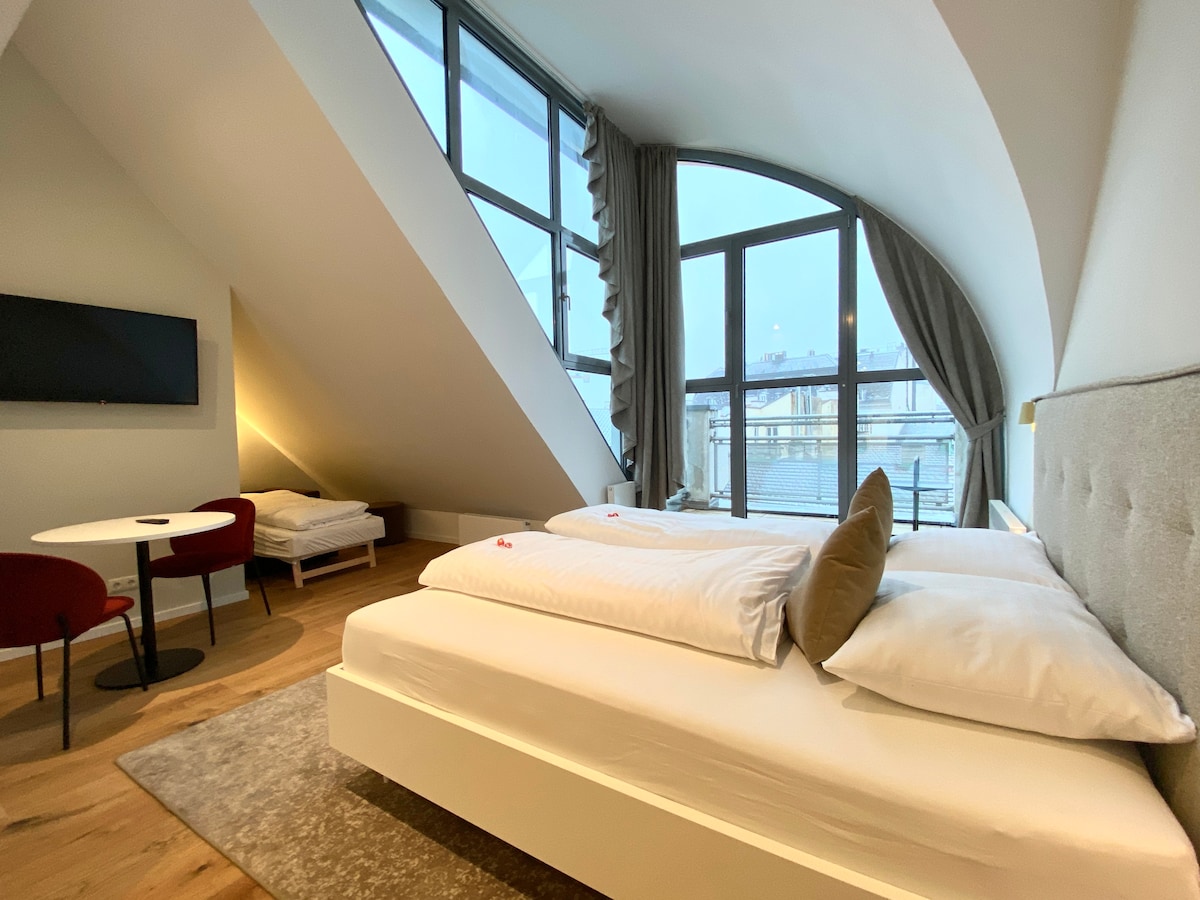 A modern room features an angled ceiling complemented by large windows that allow natural light to fill the space. A spacious bed with white bedding is visible, along with a small seating area equipped with a round table and two chairs. A single bed is positioned nearby.