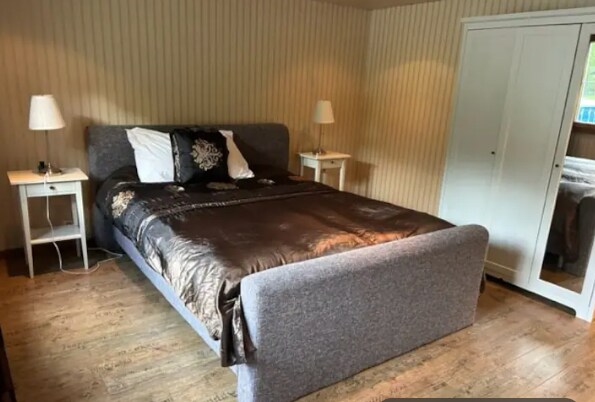 A spacious bedroom features a large upholstered bed covered with a chocolate brown duvet. Two bedside lamps are positioned on white nightstands on either side. A built-in wardrobe with mirrored doors complements the warm wood flooring and soft wallpaper.