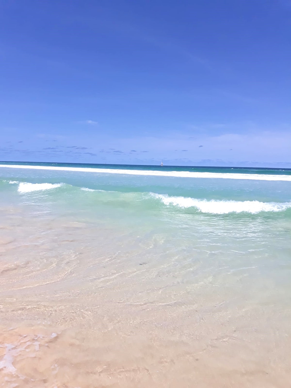 Gentle waves lap against a sandy shore, reflecting vibrant shades of blue and green. The horizon showcases a clear sky, with wispy clouds scattered above. The tranquil ocean waters create a serene atmosphere, perfect for relaxation.