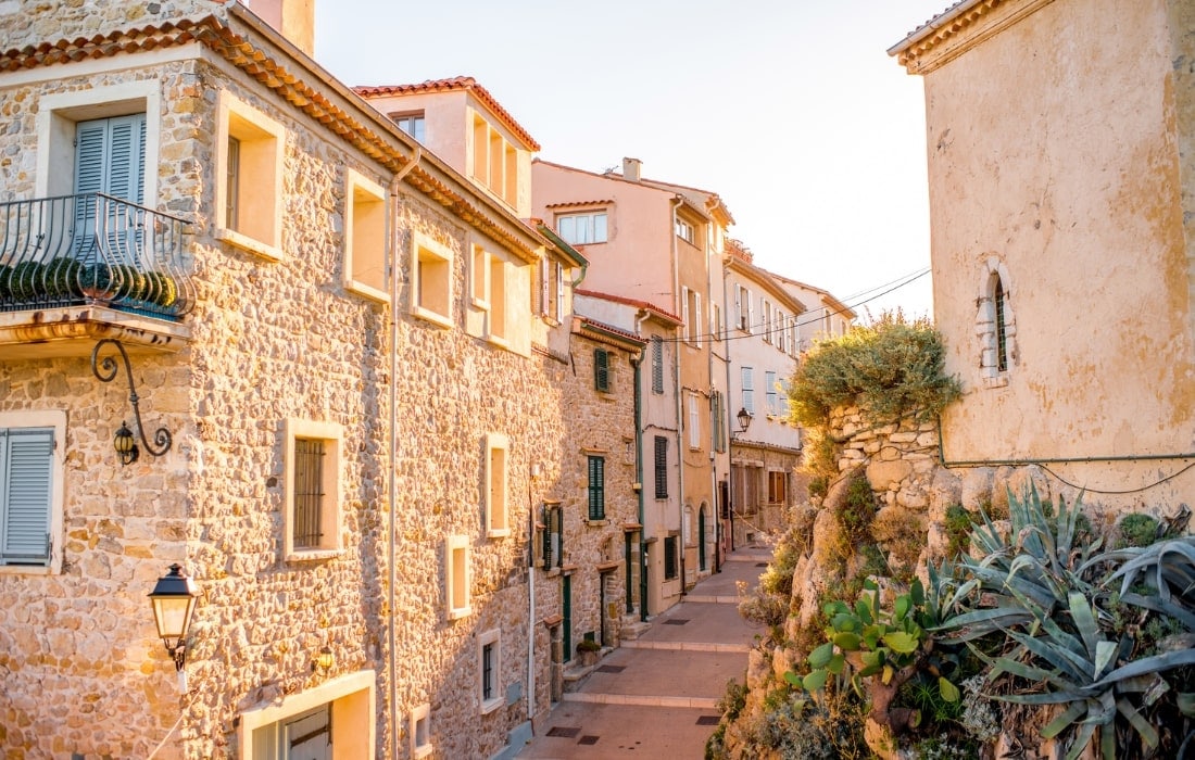 A charming street is framed by stone buildings, showcasing traditional architecture in warm hues. Soft sunlight bathes the area, highlighting the details of the facades and balconies. Lush greenery and potted plants are visible along the pathway, enhancing the inviting atmosphere of this historic locale.