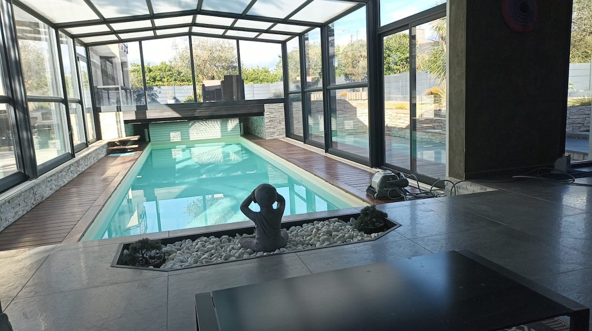 An indoor pool area is enclosed by large glass panels, allowing natural light to illuminate the space. A sculptural feature is positioned at the edge of the pool, surrounded by decorative stones. A smooth floor extends around the pool, maintaining a clean and modern aesthetic.