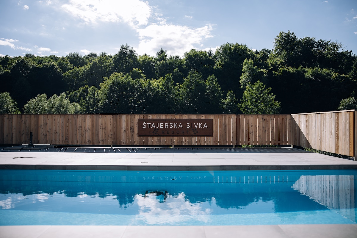 The inviting outdoor pool is bordered by a wooden fence and reflects the surrounding greenery, creating a serene atmosphere. The sign reading 'Štajerska Sivka' is prominently displayed, adding a distinct touch to the scene. Soft natural light enhances the tranquil setting.
