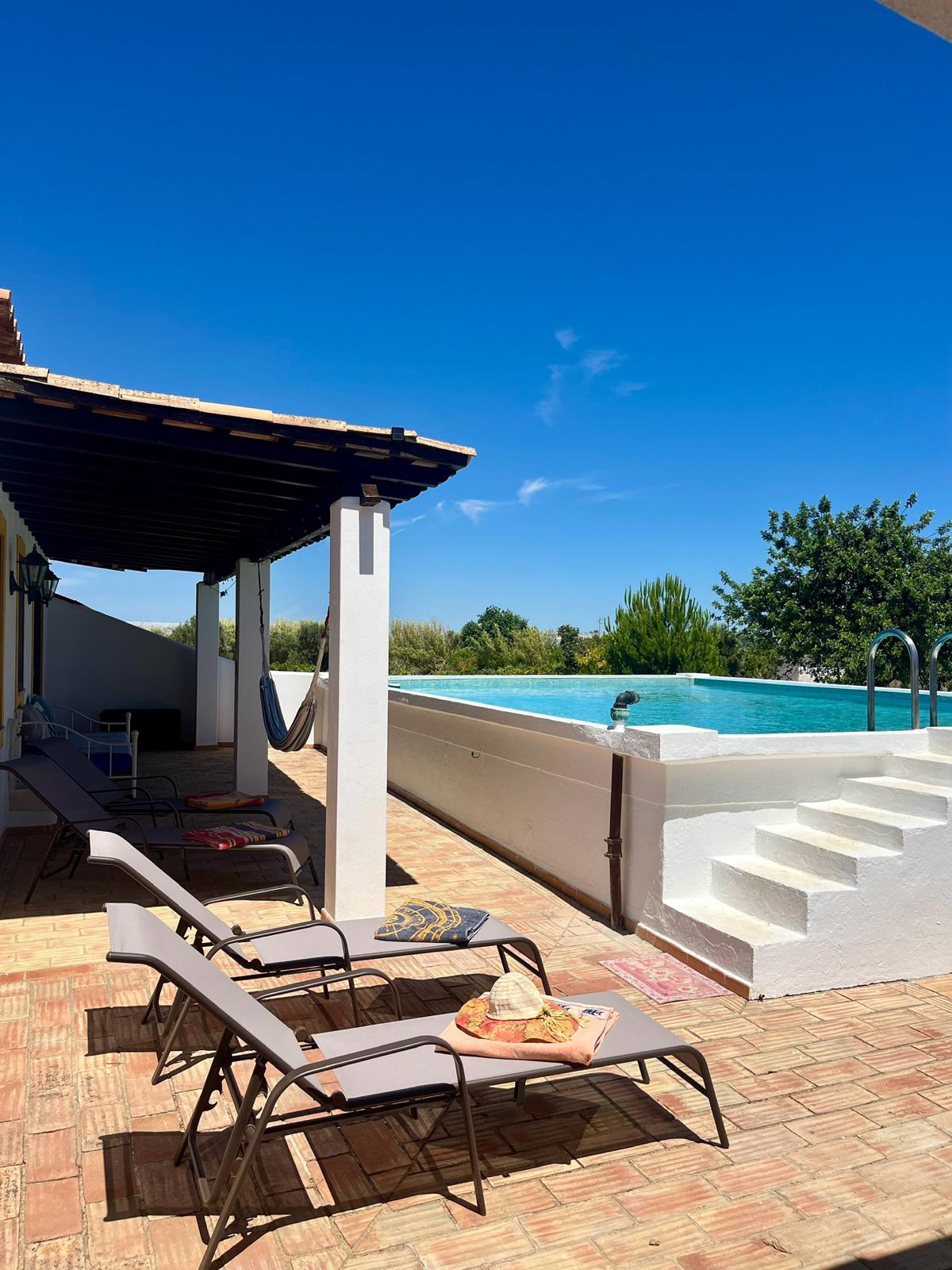 A sunlit terrace area features several lounge chairs positioned around a serene private pool. Steps lead gently into the water, while the surrounding landscape includes lush greenery and a clear blue sky overhead.