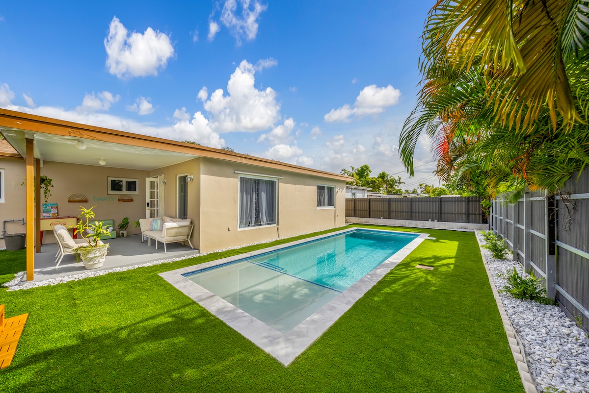 A spacious backyard is highlighted by a sleek pool, surrounded by well-maintained artificial grass. A covered patio offers comfortable seating, while sun loungers invite relaxation under the sunny sky. Lush palm trees add a touch of greenery, enhancing the serene atmosphere of the outdoor area.