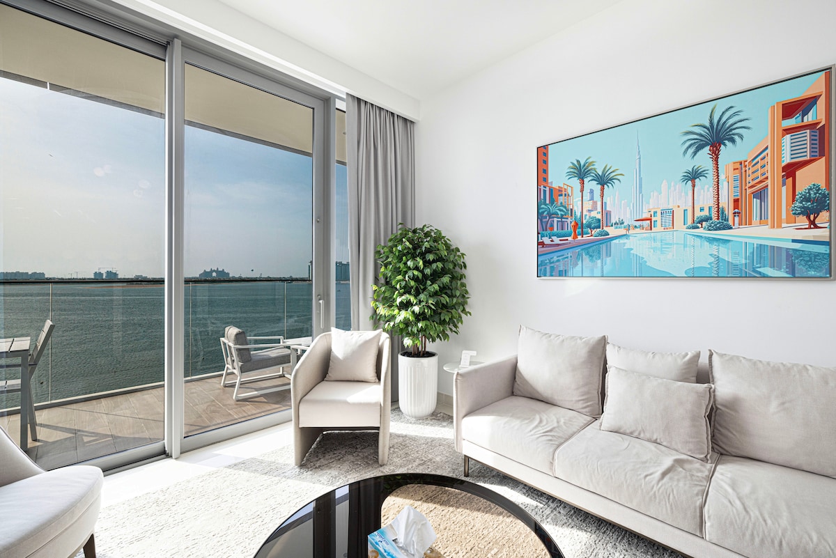 A bright living room features large glass doors that open onto a terrace, providing impressive views of the water. Elegant, neutral-toned sofas surround a black circular coffee table. A vibrant artwork complements the space, while a potted plant adds a touch of greenery.