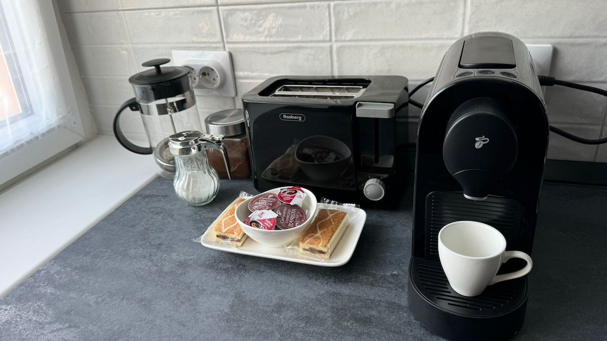 A kitchen countertop showcases a coffee maker next to a toaster and a French press. A small glass jar holds sugar, while silverware can be seen in the background. A plate with cookies and a white cup sit invitingly at the front.