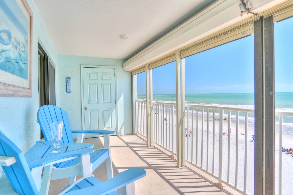 A private balcony features two blue Adirondack chairs, positioned to overlook the sandy beach and the Gulf of Mexico. The door to the condo is visible, along with a clear view of the calm ocean waters and bright blue sky.