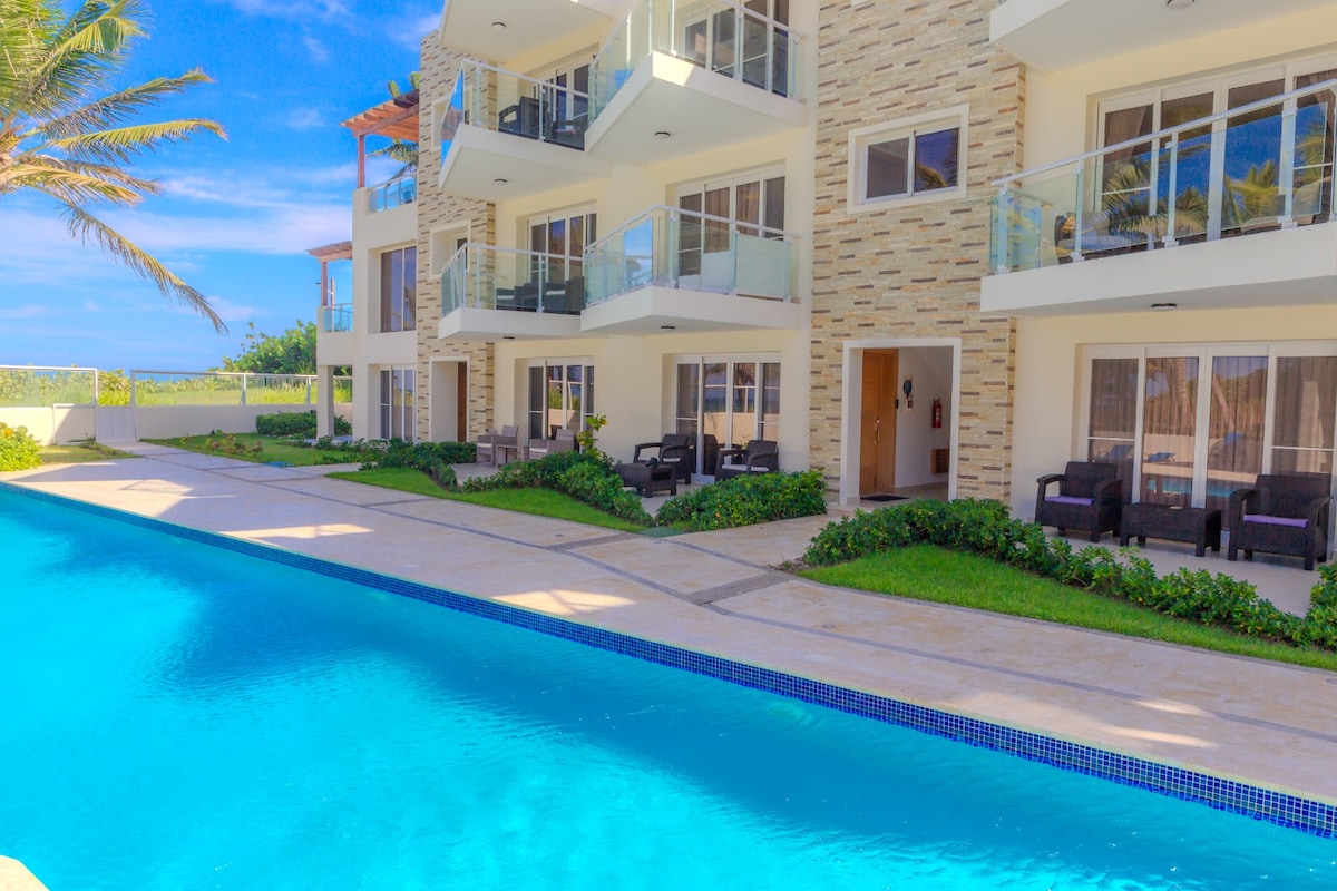 The ground floor condo, situated poolside, features a serene swimming pool with clear blue water. Spacious balconies offer a view of the pool and surrounding greenery. The modern building's facade is highlighted by large windows and a natural stone finish, creating a welcoming environment.