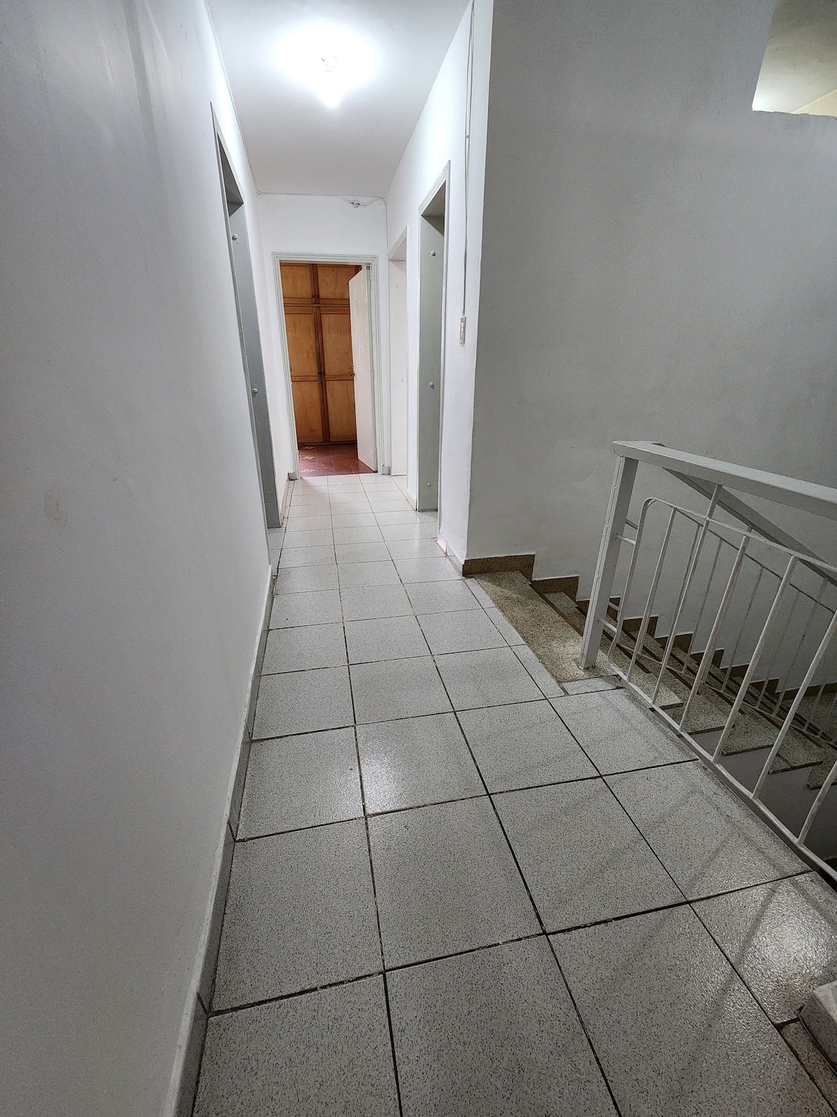 A clean and well-lit hallway is visible, featuring white walls and tiled flooring. Four doors are positioned along one side, leading to individual rooms, while a staircase is seen at the end of the corridor, offering access to lower levels.