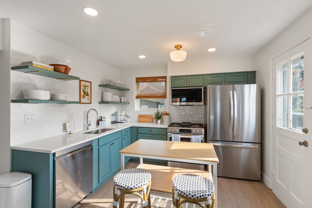 A modern kitchen is equipped with stainless steel appliances and a mix of blue and white cabinetry. A small table with striped chairs is positioned in the center. Open shelving displays dishware, while a door leads to the outside, offering natural light.
