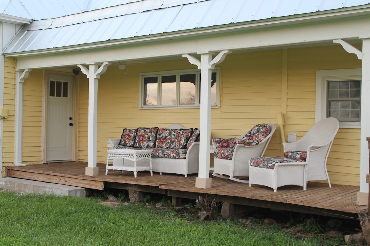 A covered porch area features multiple seating options, including a large floral-patterned sofa, a wicker rocking chair, and a small side table. The inviting yellow exterior of the house is complemented by white trim and a light blue metal roof.