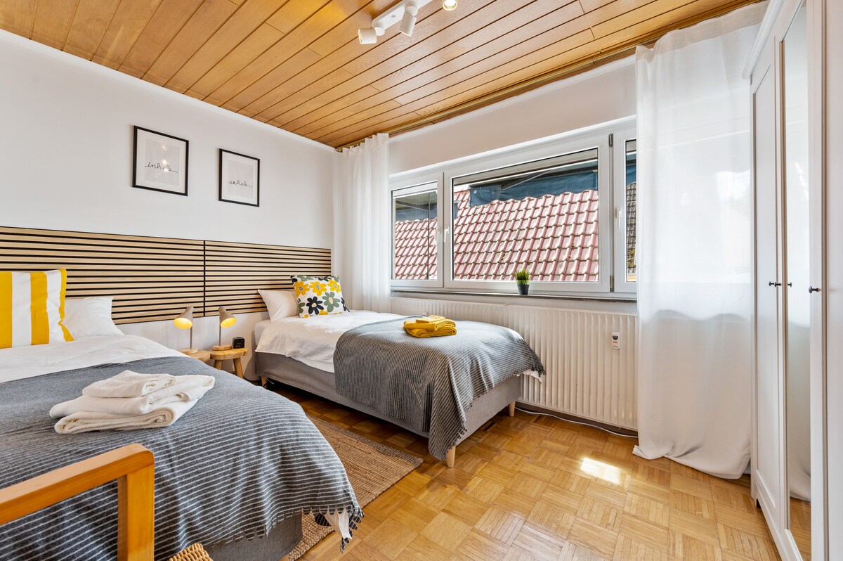 A well-lit bedroom features two comfortable beds with soft bedding, positioned near a window with sheer curtains. Light wood paneling adorns the ceiling, and wall art hangs above the beds. A stylish rug covers the parquet flooring, complemented by bedside tables.