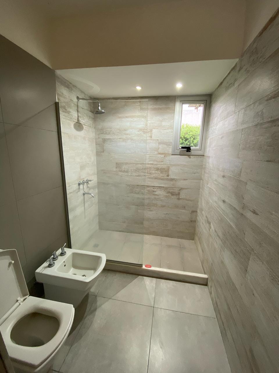 A modern bathroom is shown, featuring a shower area with a glass panel and tiled walls. A compact toilet and a bidet are positioned adjacent to each other. A small window allows natural light to enter, and the floor is tiled in a neutral grey tone.