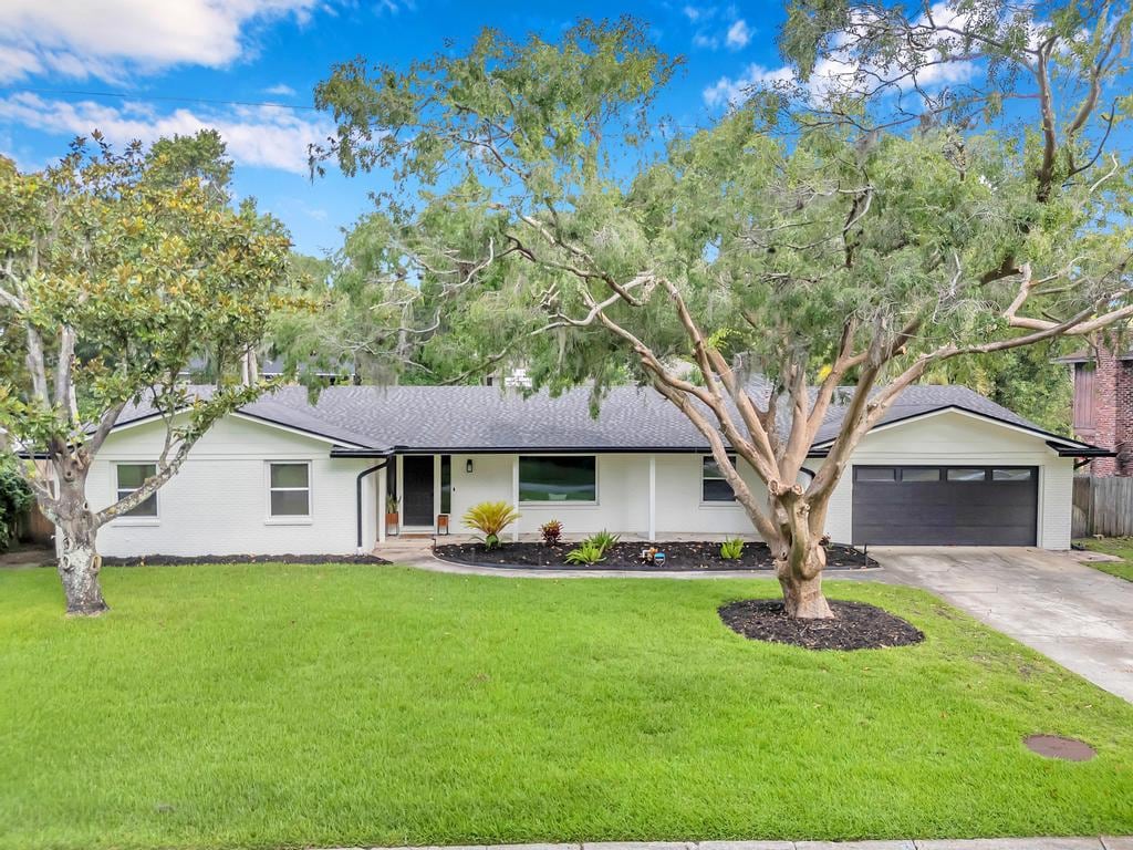 The exterior of a single-level home is shown, featuring a manicured lawn and two large trees in the front yard. A concrete driveway leads up to a garage, while colorful plants are neatly arranged near the entrance path, creating a welcoming appeal.