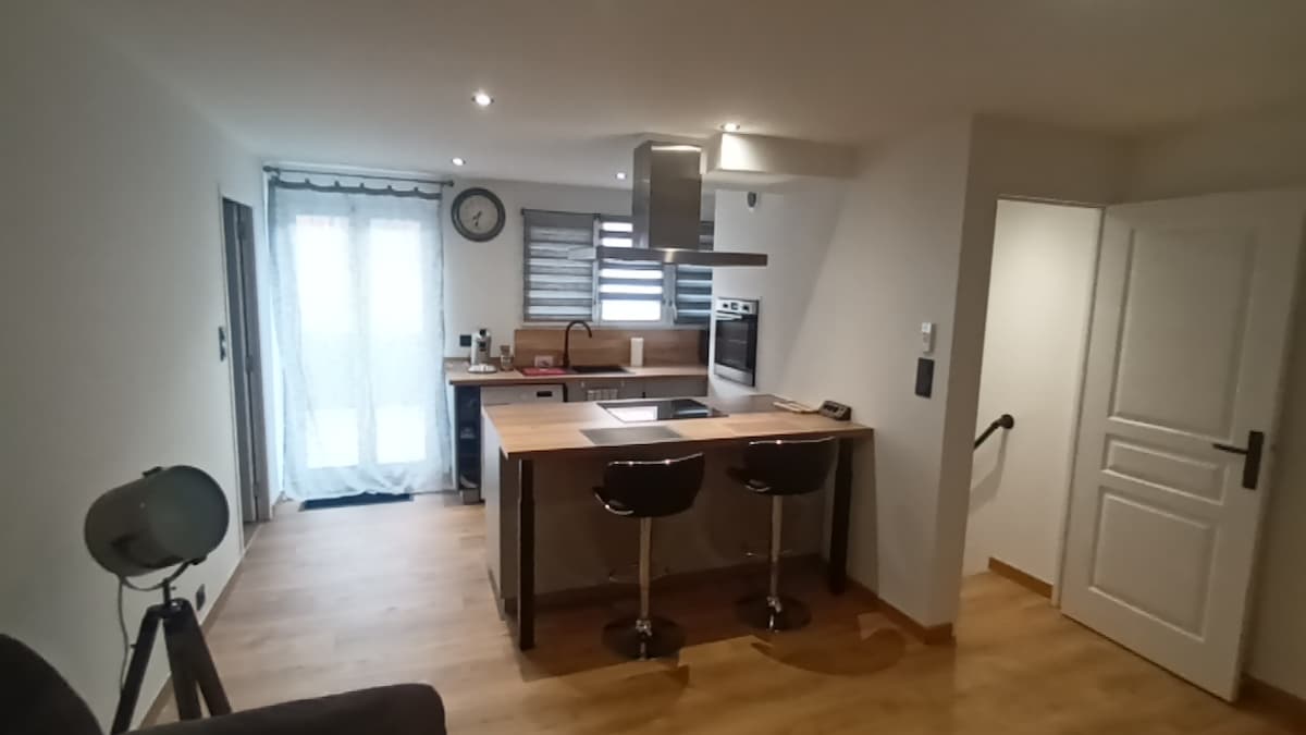 The kitchen features a modern design with wooden countertops and sleek appliances. A stylish island with two bar stools is positioned at the center. Natural light enters through a sheer curtain on the window, illuminating the space. The area includes essential kitchen equipment and storage.