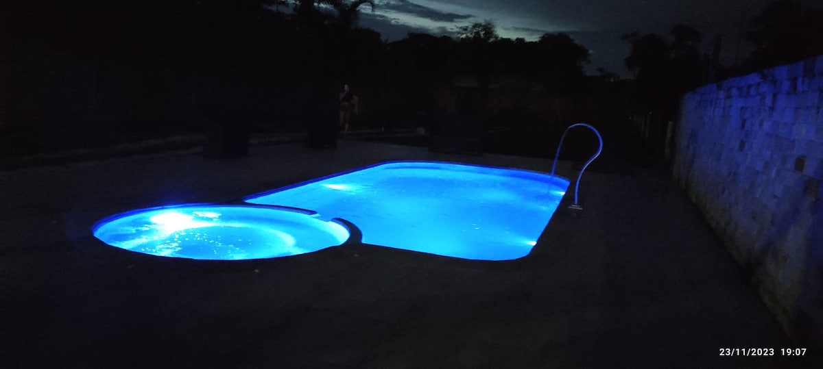 A pool area is illuminated by blue LED lights at night, highlighting the inviting shapes of the main pool and an attached hot tub. The surrounding space is dark, creating a serene environment for relaxation.