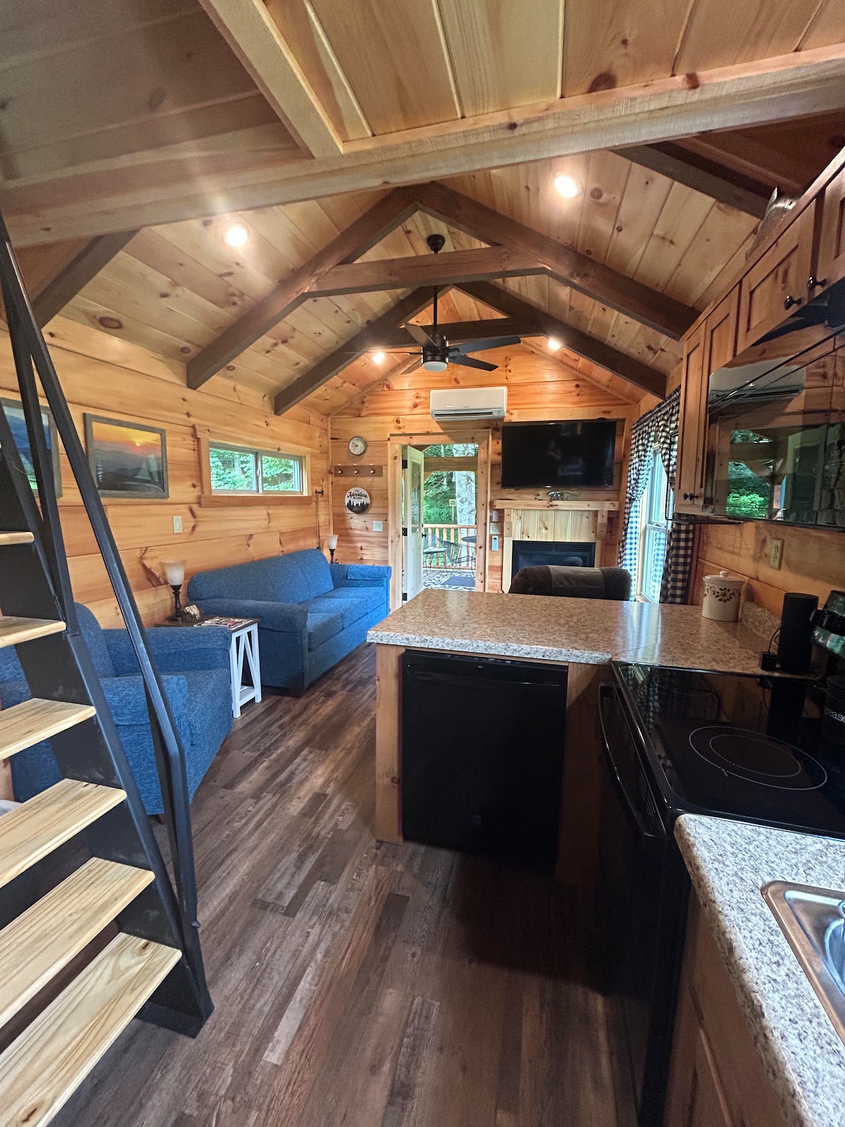 The interior features wooden walls and ceiling, creating a cozy ambiance. A spacious kitchen area with granite countertops is visible, alongside a comfortable seating area with blue couches. Large windows allow natural light to enhance the open layout, leading to a door that opens to the outdoors.