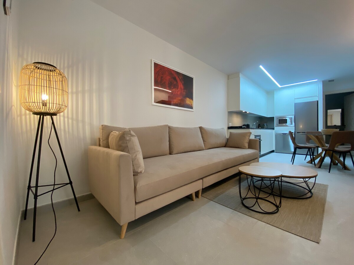 A cozy living area features a light beige sofa complemented by a round coffee table with a wireframe design. A modern floor lamp provides warm light, and a framed artwork adds a touch of color to the neutral walls. The open kitchen area can be seen in the background.