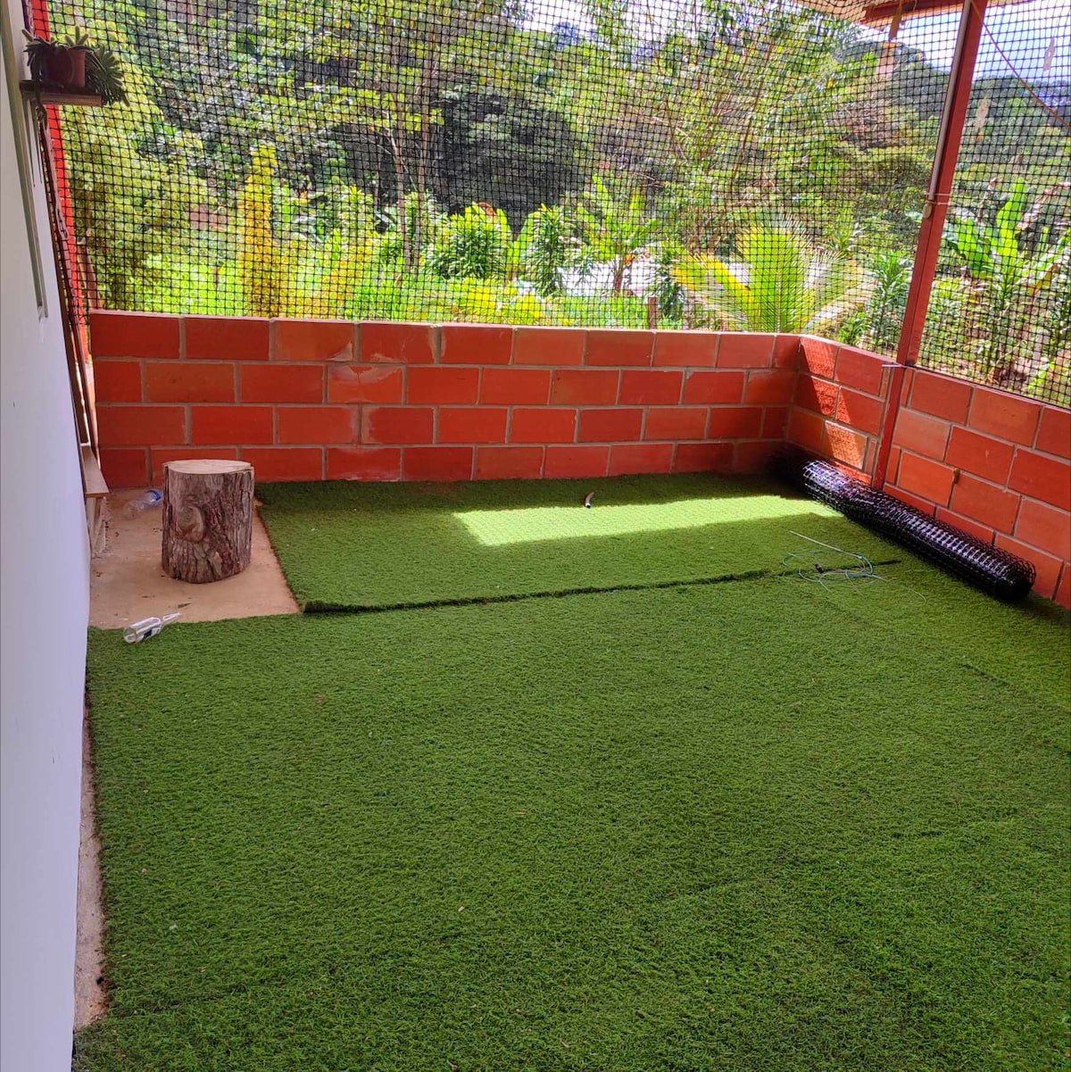An outdoor area is displayed with artificial grass covering the ground, bordered by a red brick wall. A small log is positioned on one side, and a roll of synthetic turf is placed along the opposite wall. Natural greenery rises in the background.