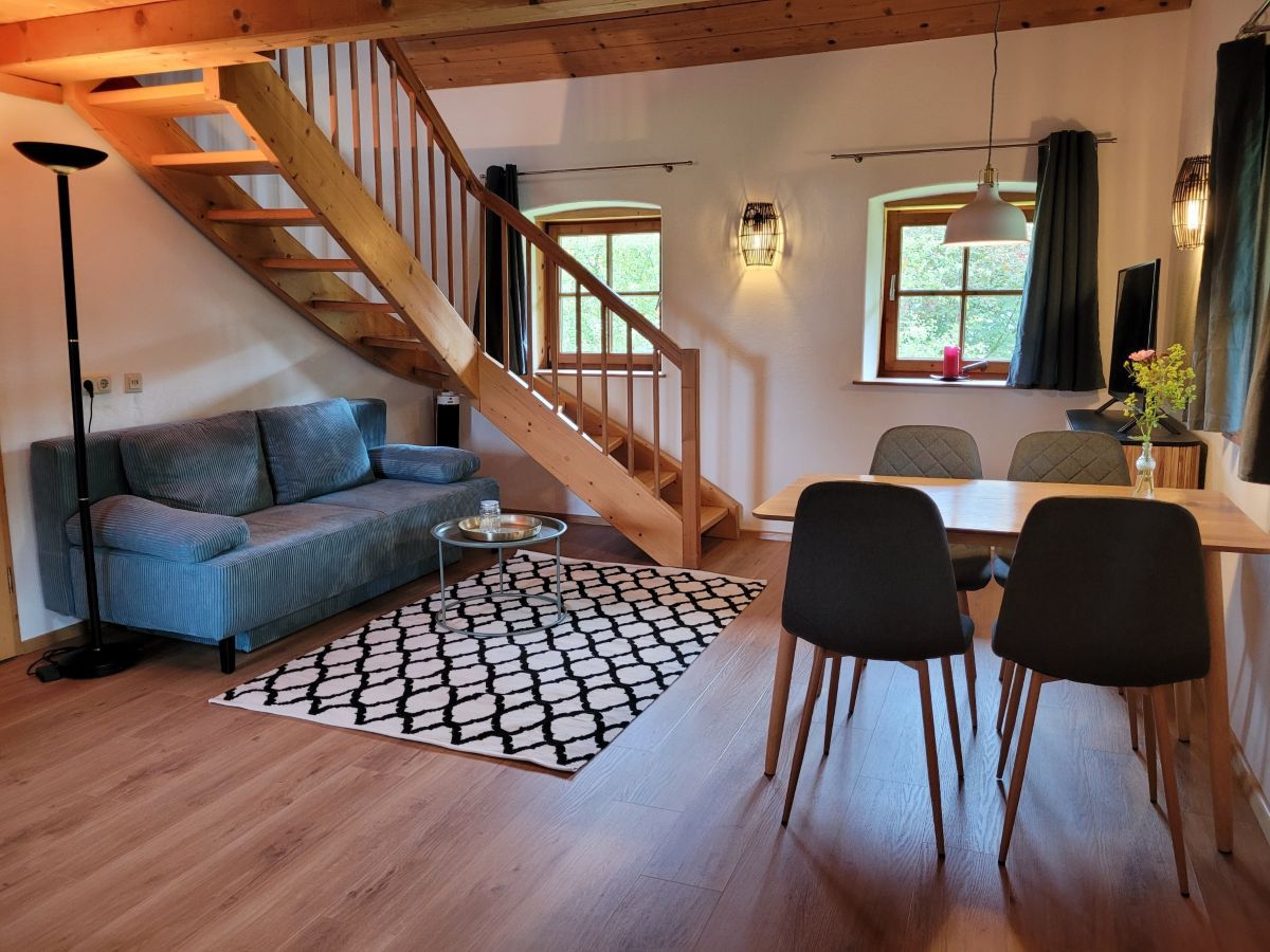 A clear view of the open living and dining area is presented, featuring a comfortable sofa beside a staircase. A dining table with two chairs is positioned nearby, complemented by a decorative rug. Natural light enters through two windows, enhancing the inviting space.