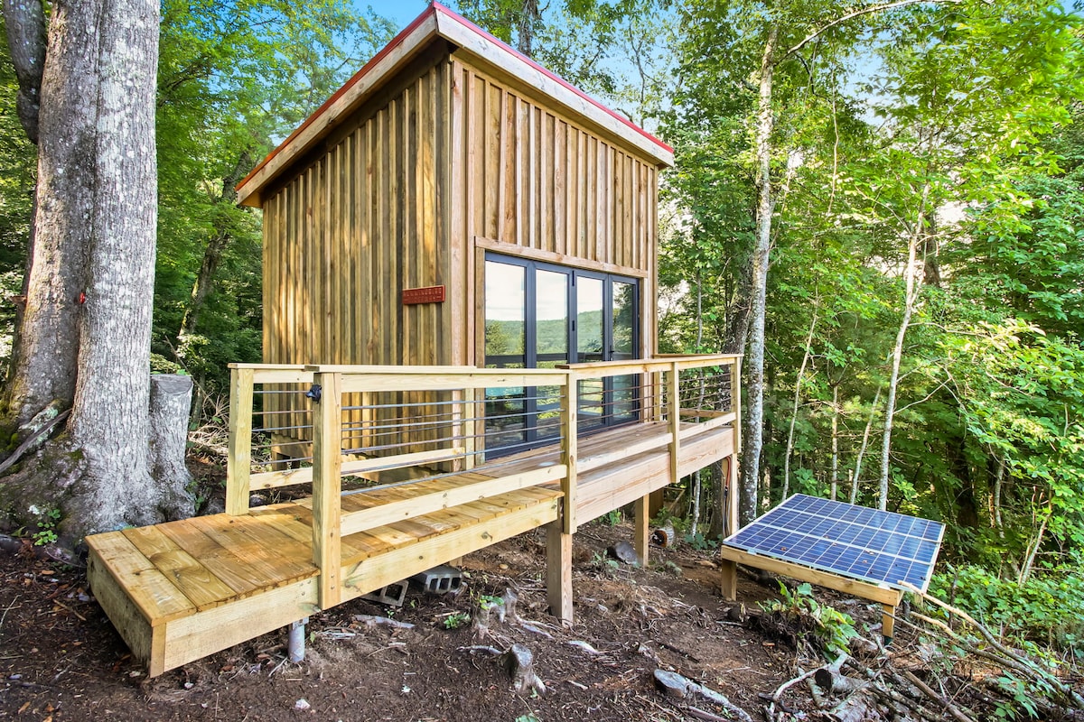 A rustic cabin features a porch with a wooden railing, leading to large glass doors that highlight the natural surroundings. A solar panel is situated beside the cabin, providing sustainable energy. Lush greenery envelops the building, enhancing its secluded atmosphere.