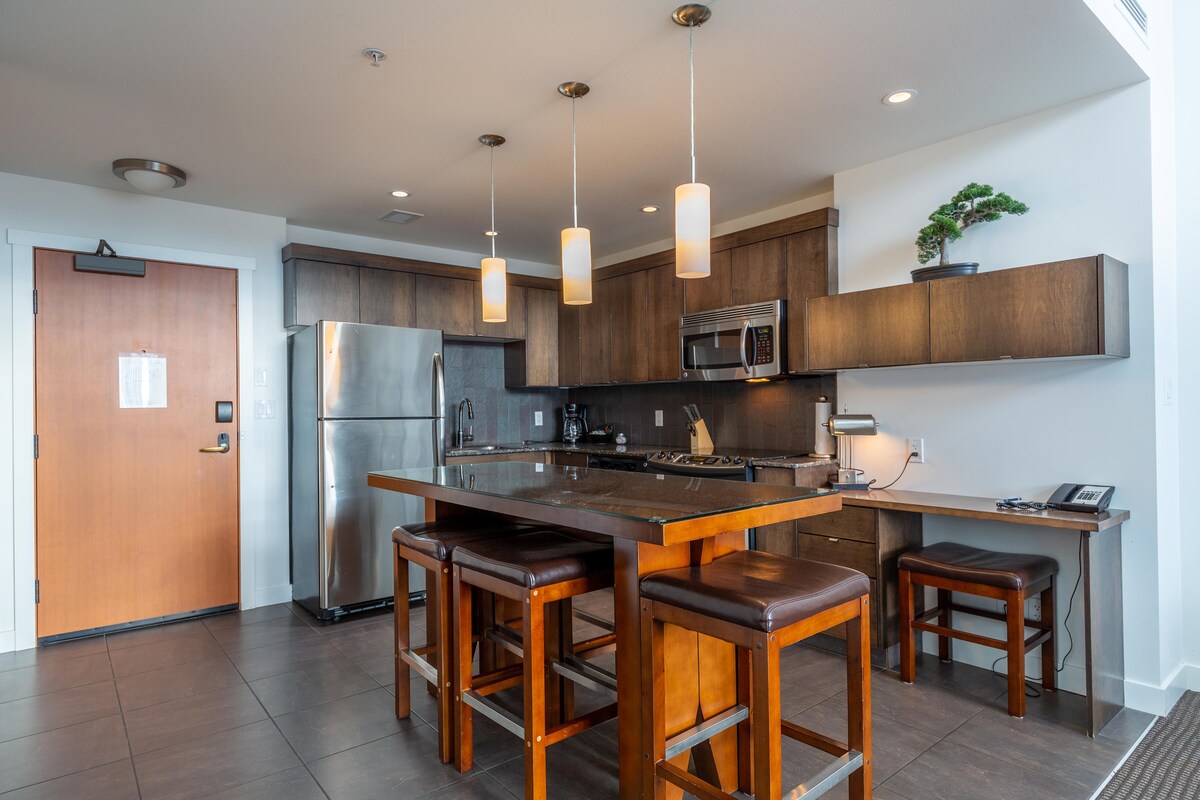 A modern kitchen space is equipped with stainless steel appliances and wooden cabinetry. An island with high stools provides seating, while pendant lights illuminate the area. A door on the left leads to the hallway, adding functionality to the layout.