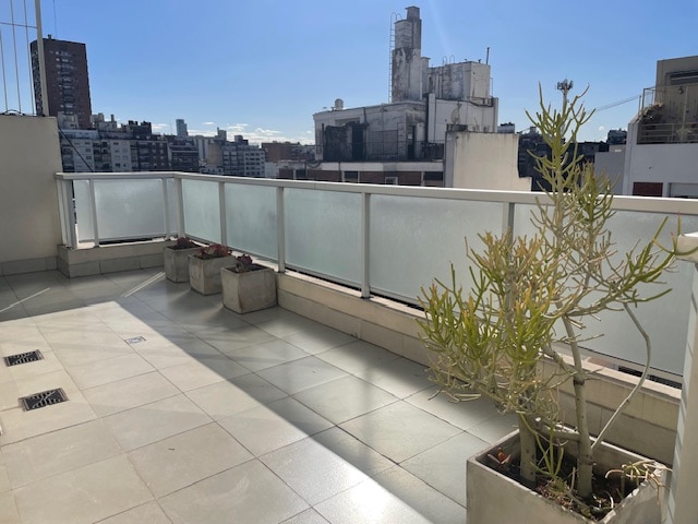 A sunlit terrace features a smooth tiled floor and a railing that provides views of the urban landscape. Several planters containing greenery are positioned along the edge, enhancing the outdoor area. The backdrop includes city buildings and a clear blue sky.