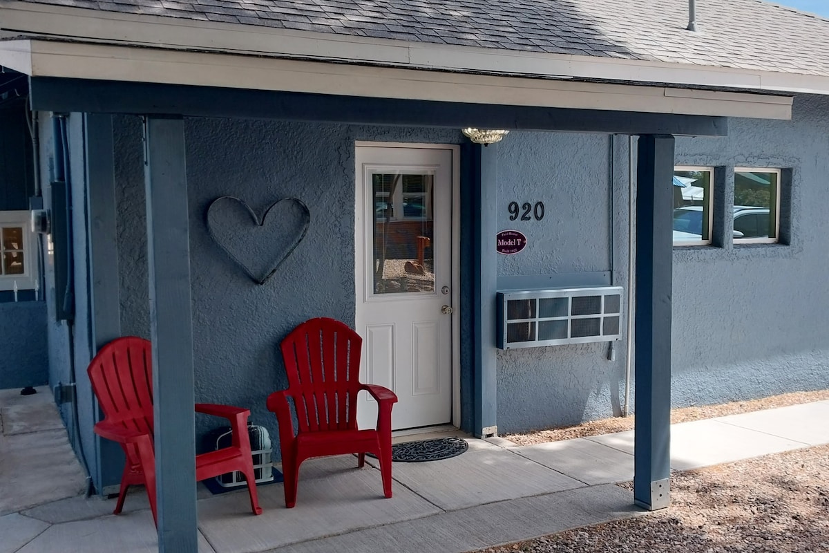 The exterior of the duplex home features a welcoming entryway with a light blue facade. Two red chairs are positioned under a covered porch next to the door. A heart-shaped decoration is mounted on the wall, and a mailbox is visible beside the entrance.