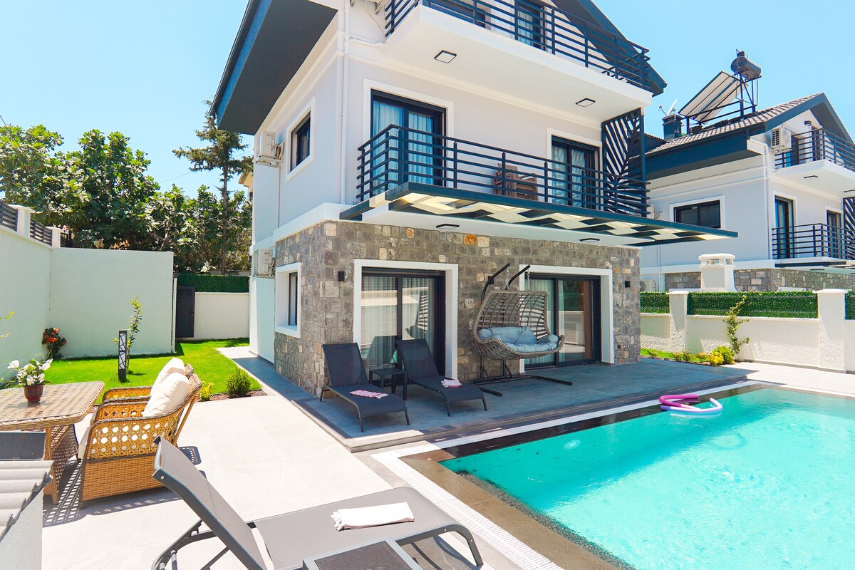 A well-maintained outdoor area features a private pool surrounded by lounge chairs and a shaded seating area. The villa's modern façade is accented by stone and is complemented by neatly trimmed greenery, creating a functional space for relaxation and enjoyment.