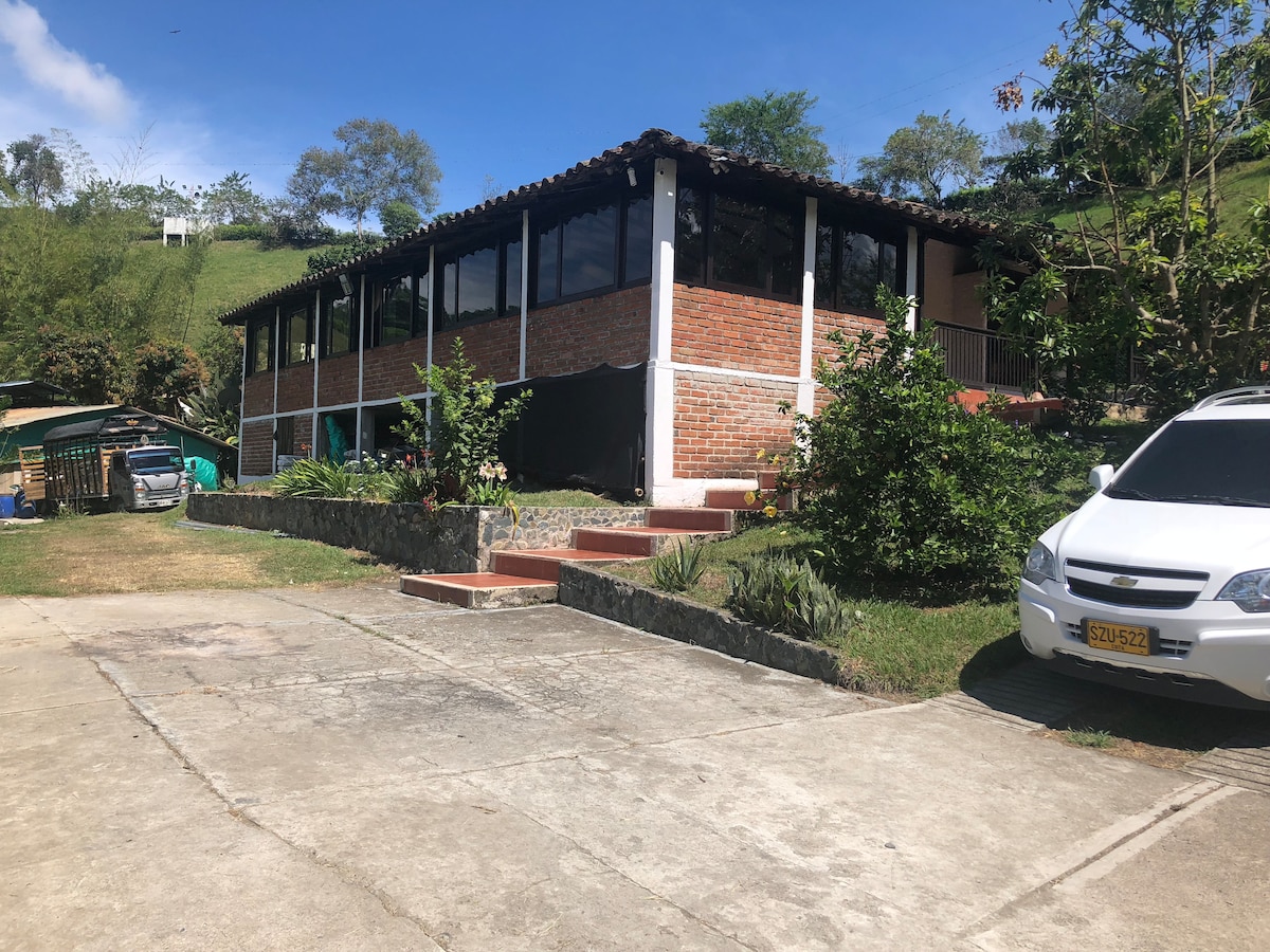A charming single-story building is set against a hillside, featuring a brick facade and spacious glass windows. Greenery surrounds the structure, with a paved walkway leading to the entrance. A vehicle is parked nearby, complementing the natural environment.