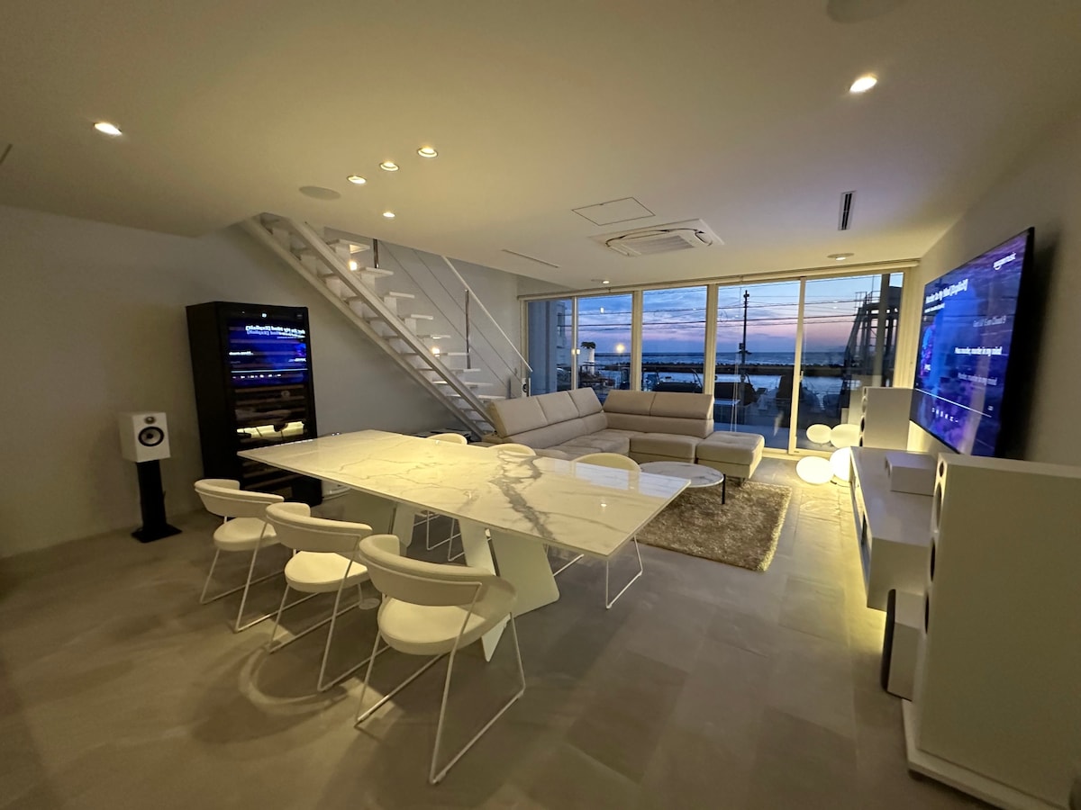 A modern living and dining area features a sleek marble dining table surrounded by white chairs. A comfortable sofa is positioned nearby, with large windows offering expansive views. A substantial flat-screen television is mounted on the wall, enhancing the contemporary ambiance.