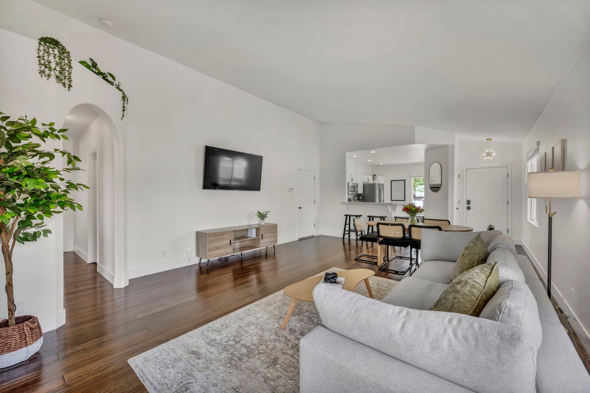 A modern living area showcases a light-colored sectional sofa, complemented by a coffee table. A flat-screen television is mounted on the wall, enhancing the spaciousness. Natural light fills the room through nearby windows, and decorative plants add touches of greenery.