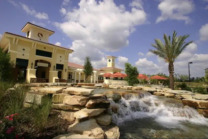 A resort entrance is showcased with a charming waterfall flowing over natural stone. Lush greenery surrounds the area, while a palm tree stands in the foreground. The building features a distinctive clock tower and outdoor seating with shaded umbrellas, complemented by a blue sky with scattered clouds.